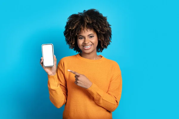 不错的移动应用程序 可爱而快乐的年轻黑人女士 留着浓密的头发和牙套 指着手机 手里拿着白色的空白屏幕 微笑着 蓝色背景 — 图库照片