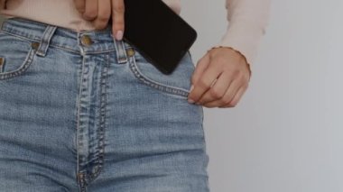 Modern hayatın aksesuarları. Tanınmayacak kadar zayıf bir kadının akıllı telefonunu kot pantolonun, yavaş çekimin, boş bir alanın cebine koyarkenki yakın çekimi.