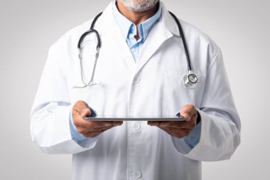 Steteskoplu beyaz önlüklü yaşlı Avrupalı erkek doktor gri stüdyo arka planında tablet tutuyor, kırpılmış. Cihaz, sağlık hizmetleri uygulaması, ilaç servisi, hastalık tedavisi ve danışma