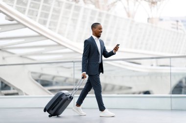 Havaalanında bavulla yürüyen yakışıklı siyahi işadamı ve akıllı telefon kullanan, takım elbiseli, cep telefonu takan ve uçağa binerken mesajlaşan Afro-Amerikan erkeği.