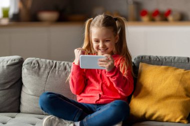 Evde akıllı telefonda oyun oynayan, başarıyı kutlayan, neşeli, genç bir kızın yumruk sallaması ve evet demesi, oturma odasında otururken online kazanmanın keyfini çıkarması.
