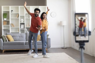 Evde video çekerken mutlu Hintli genç çift dans ediyor, pozitif doğu eşleri sosyal ağlar için içerik yaratıyor, oturma odasında tripodda akıllı telefon kullanıyor, boşluğu kopyalıyor.