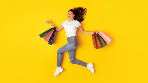 购物和销售 愉快的女士采购跳跃携带许多纸袋投递在工作室黄色背景 女性购物狂对相机微笑广告季节性折扣 — 图库照片