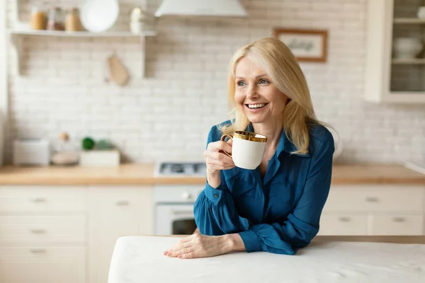 キッチンだけで新鮮なコーヒーや紅茶を飲む魅力的なブロンドの成熟した女性は 脇を見て笑顔で 無料のスペース 笑顔の女性がカップを保持し 居心地の良いキッチンに立って — ストック写真
