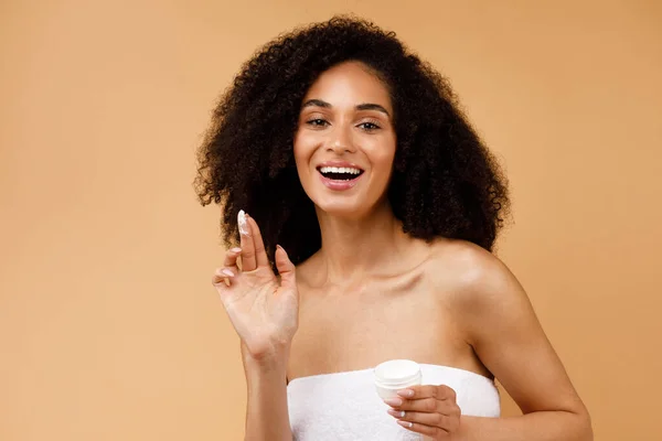 スキンケアの概念 かなりアフリカ系アメリカ人女性保湿クリームを適用します タオルで包まれたポーズとカメラで笑顔 茶色の背景の上に立って — ストック写真