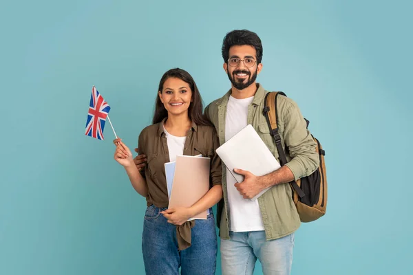快乐而美丽的多民族千禧年夫妇阿拉伯裔印度男生 笔记本电脑和记事本 展示英国国旗 出国留学 学习英语 有蓝色工作室背景 — 图库照片