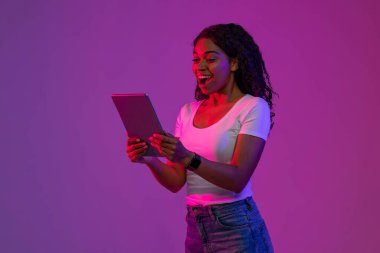Heyecanlı genç Afrikalı Amerikalı Kadın Dijital Tablet Tutuyor ve Ağzı Şaşırarak Açılıyor, Mor Stüdyo Arkaplanının Üzerinde Neon Işığı Altında Dururken Modern Gadget kullanan Siyah Kadın Şaşırdı