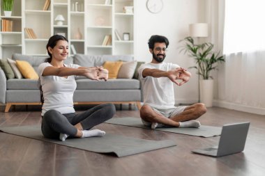 Gülümseyen Hintli çift dizüstü bilgisayarın önünde yan yana sırıtıyor, mutlu doğu eşleri evde antrenman yapıyor, ev içi spor yapıyor, oturma odasında spor yapıyor, boş alanda spor yapıyor.