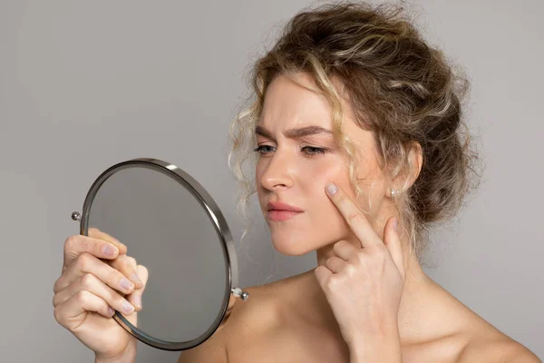 Upset lady holding magnifying mirror and looking at pimple on cheek, frustrated woman having acne on face, suffering problem skin, standing on grey studio background