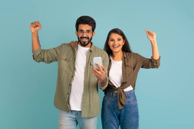 Duygusal olarak mutlu genç Hintli çift online kumar oynuyor, mavi stüdyo arka planında telefon kullanıyor, yumruklarını kaldırıyor ve başarıyı kutluyor. Çevrimiçi bahis, alım satım, nakde çevirme, dağıtma