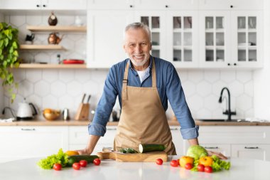 Sağlıklı yemek pişirirken mutfakta poz veren yakışıklı yaşlı adam, önlük giyen mutlu yaşlı beyefendi evde sebze yemeği hazırlayan yaşlı şef kameraya gülümsüyor, mekanı kopyalıyor.