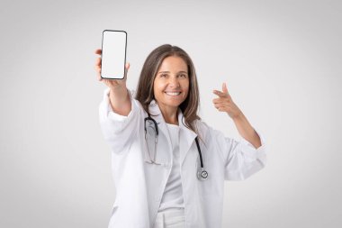 Neşeli kıdemli doktor kadın cep telefonunu boş ekranla gösteriyor, gri arka planda duruyor, tıbbi uygulama için maket yapıyor. Pratisyen hekim telefonu işaret ediyor.