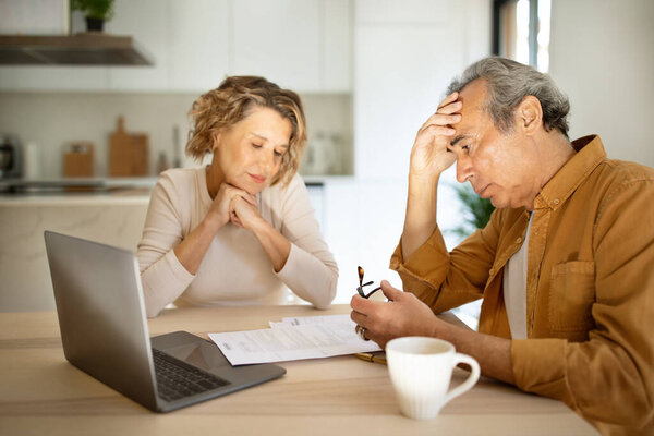 Stressed elderly couple checking financial papers, sitting with laptop and looking at loan documents, suffering crisis while calculating family spends, kitchen interior