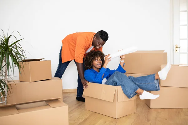 Happy African American Spouses Moving Home And Having Fun Together, African American Spouses Fooling While Packing Things On Relocation Day, Man Pushing Cardboard Box With His Happy Wife