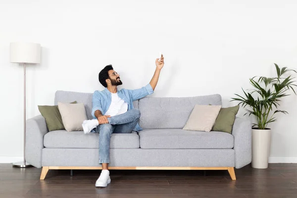 stock image Happy handsome bearded middle eastern young man in casual outfit sitting on couch at home, holding remote control, turning on ac, pointing towards copy space on white wall