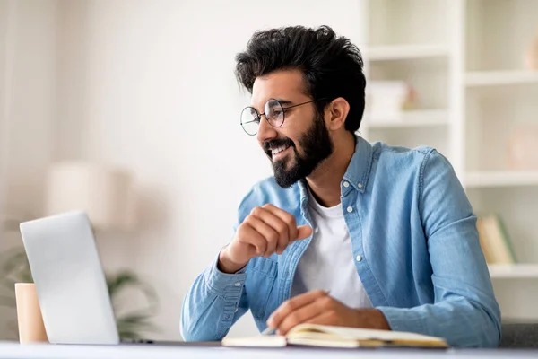 Young indian man using laptop and taking notes while working at home office, smiling eastern male freelancer sitting at desk, looking at computer screen and writing in notepad, copy space