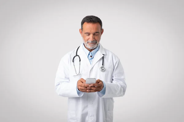灰色のスタジオの背景に電話でチャット聴診器と白いコートの深刻な古いヨーロッパの男性医師 ヘルスケア 医療サービス 疾患治療 遠隔相談のためのアプリ — ストック写真
