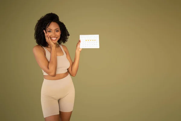 穿着运动服的快乐的年轻非洲裔美国卷曲纤细女士在米色工作室背景下展示日历 适合的生活方式 身体护理 积极锻炼 减肥时间表 — 图库照片
