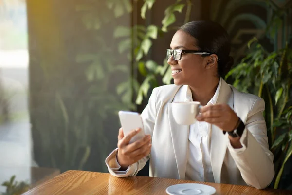 Cheerful young black woman in suit with glasses at table, looks at window at copy space with phone in cafe with plants interior. Rest, coffee break, device for modern business and work, create idea