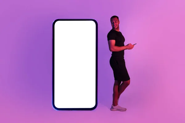スポーツ用途 スタジオで紫ネオンの背景に空の画面を持つ大規模なスマートフォンの近くに立って電話を使用してアフリカ系アメリカ人スポーツマン デジタルワークアウトを提供します モックアップ — ストック写真