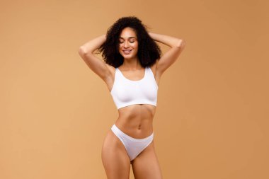 Beyaz iç çamaşırlarıyla poz veren, bej renkli stüdyo arka planında elleri ve gözleri kapalı seksi Afro-Amerikalı kadın.