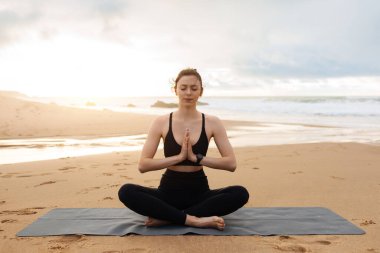 Spor giyim bölümünde yoga yapan sakin bir kadın okyanus kıyısında, gözleri kapalı meditasyon yapıyor, fotokopi çekiyor. Spor, kilo kaybı ve açık havada antrenman.