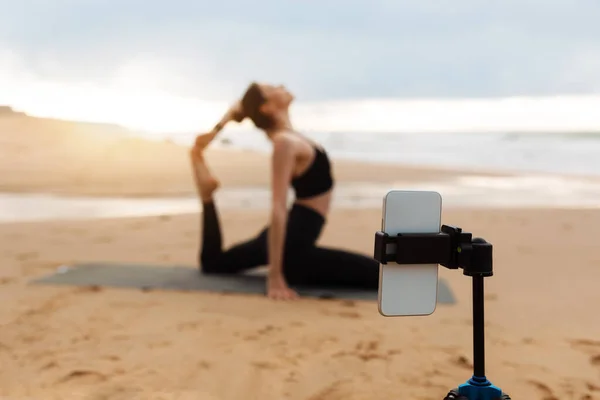 Female yoga teacher leading online lesson on beach by the ocean, broadcasting to camera of smartphone standing on tripod, selective focus. Blogging, vlog. Healthy lifestyle
