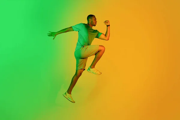 坚定的非洲裔美国跑步者盖伊跳跃 在黄色工作室背景下的绿色霓虹灯下练习 适合运动员跑向后看 侧视图 全长镜头 — 图库照片