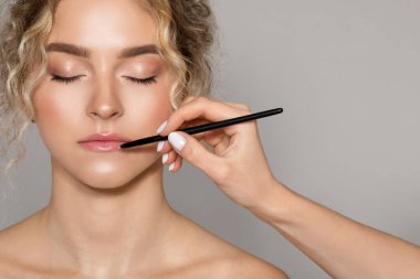 Profesyonel makyaj sanatçısı, beyaz kadın için çıplak makyaj yapıyor, gri stüdyo arkaplanı üzerine fırçayla ruj sürüyor, yakın plan, kopyalama alanı