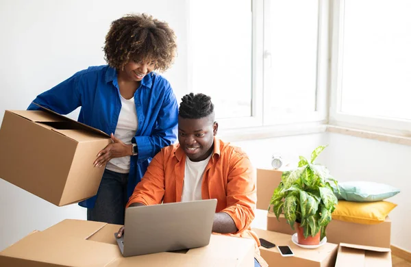 Neşeli Siyah Çift Laptop Kullanıyor, Taşınan Şirketleri Çevrimiçi Kontrol Ediyor, Mutlu Afrikalı Amerikalı eşler Karton Kutular Arasında Otururken Bilgisayar Kullanıyor, Boşluğu Kopyala