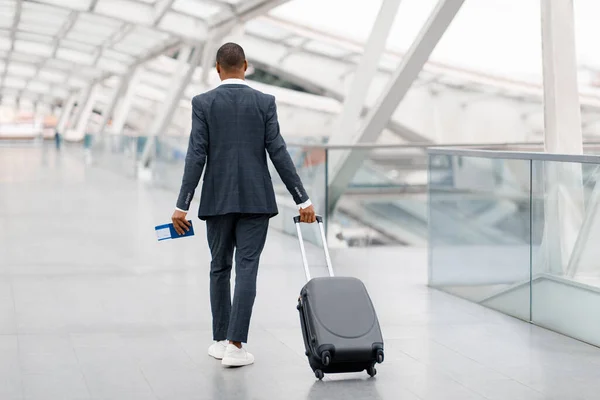 Black Man In Suit Walking With Suitcase At Airport Terminal, Real View Of African American Businessman Carrying Luggage And Holding Passport With Tickets While Going To Flight Gate, Copy Space