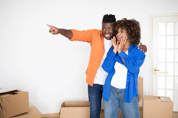 Excited black man surprising his wife with moving to new home, young african american family couple standing in apartment among carton boxes, cheerful spouses celebrating first day in their own house