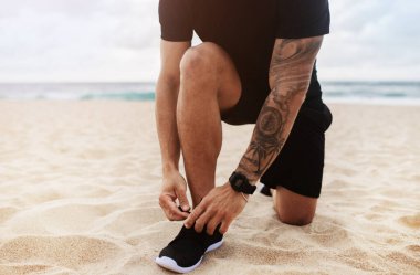 Spor kıyafetli bir adam okyanus sahilinde koşarken ayakkabılarını bağlıyor, bacaklarını kapatıyor, manzarası kesiliyor. Genç erkek dışarıda koşuyor, sağlıklı aktif yaşam tarzının tadını çıkarıyor.