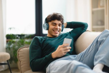 Orta Doğulu gülümseyen, akıllı telefon kullanan, kulaklık takan ve evdeki koltukta uzanmış müzik dinleyen bir adam. Genç Adam Haftasonunun tadını çıkartıyor. Özellikler ve İnternet
