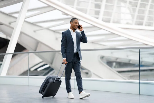 携帯電話だ ターミナルでスーツケースを持って歩いている間 空港で携帯電話で話している若い黒人ビジネスマンの笑顔 快適な電話会話を持つスーツを着ている幸せなアフリカ系アメリカ人男性 — ストック写真