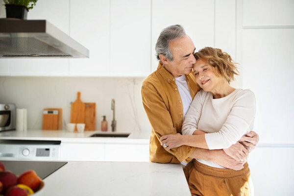 Loving senior spouses embracing, enjoying tender moments together at home, standing in kitchen interior, copy space. Romantic relationship in older age, retirement lifestyle