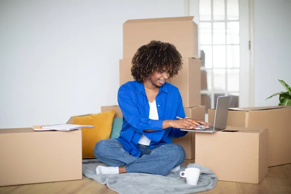 Happy Black Woman Checking Design Website On Laptop After Moving Home, Smiling African American Female Using Computer And Planning Decorations In New Apartment, Sitting Among Cardboard Boxes
