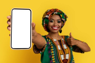 Beyaz beyaz ekranlı, güzel siyah kadın eli ile akıllı telefon, ulusal Afrika kostümü giyen mutlu genç bayan harika bir online anlaşma ve başparmak yukarı, sarı stüdyo arka planı, maket