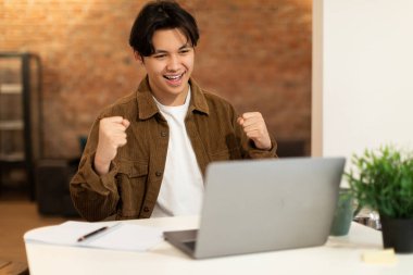 E-Öğrenme Başarı. Bilgisayarın başındaki neşeli Japon öğrenci şans ve zafer kutlaması için yumruklarını sallıyor ve evinde oturuyor. Başarılı Çevrimiçi Eğitim ve Evde Eğitim