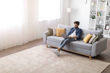 Evdeki rahat koltukta oturan genç, yakışıklı Hintli adamın portresi. İnternette bilgisayar kullanan mutlu doğu çocuğu.