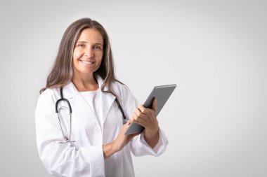 Kendine güvenen, tabletli yaşlı doktor kameraya gülümseyen, gri stüdyo arka planında poz veren, fotokopi çeken. Sağlık görevlisi, sağlık hizmetleri, dijital aygıt ve uzaktan konsültasyon için yeni uygulama