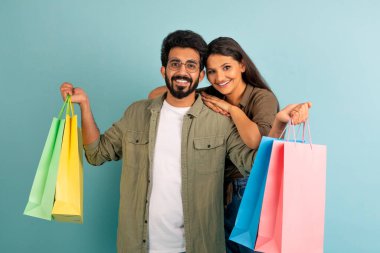 Neşeli genç Hintli çift mavi stüdyo arka planında renkli alışveriş çantalarıyla birlikte poz veriyorlar, alışverişlerini gösteriyorlar, Kara Cuma satışında alışverişin keyfini çıkarıyorlar.