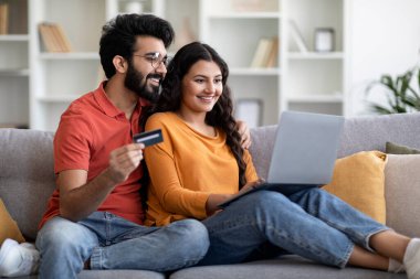 Kolay ödemeler. Laptoplu ve kredi kartlı mutlu Hintli çift evdeki koltukta dinleniyor, neşeli Doğu eşleri online alışveriş yapıyor, internetten alışveriş yapıyor, internetten alışveriş yapıyor,