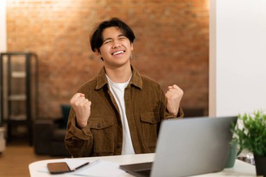 Dizüstü bilgisayarın yanında oturan neşeli Asyalı öğrenci. E-öğrenme başarısını kutluyor. Evdeki masada oturuyor. Mutlu Gençler Zaferini Kutluyor, Çevrimiçi Oyunlar Oynuyor ve Öğreniyor