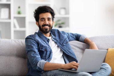 Evdeki koltukta otururken dizüstü bilgisayar kullanan yakışıklı Hintli erkek portresi, Millennial East 'li gözlüklü adam oturma odasında bilgisayarla uzaktan çalışıyor ve kameraya gülümsüyor.