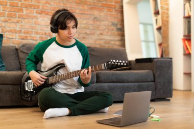 Yetenekli Koreli genç adam yeni akorlar öğreniyor dizüstü bilgisayar kullanarak evde oturuyor, kablosuz kulaklık takıyor. Teknoloji ve Müzik Konsepti