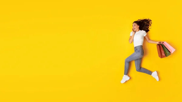 买东西 兴奋的女人提着五颜六色的纸袋 在黄色的工作室背景下在半空中奔跑 短视的女士在寻找文本的自由空间 — 图库照片