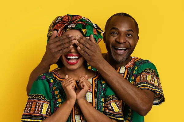 Loving handsome middle aged black man make surprise for his wife, closing excited lady eyes, couple wearing african costumes, yellow studio background, celebrating anniversary or birthday party