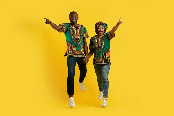 興奮した感情的なアフリカのカップルは 手を保持する伝統的な衣装を身に着けています 実行してカメラを指して 大きな取引を追いかけて 黄色のスタジオの背景 フル長さ コピースペース — ストック写真