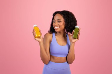 Spor giyimli neşeli Afro-Amerikan kıvırcık kadın pembe stüdyo arka planında izole edilmiş smoothie şişelerini gösteriyor. Formda yaşam tarzı, vücut bakımı, diyet ve sağlıklı içecek, süper yiyecekler ve vitaminler
